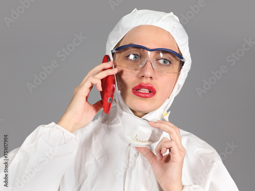 Headshot of woman in white protective suit of synthetic paper, glasses and respiritory half mask, talking on phone, disquieted, on gray background photo