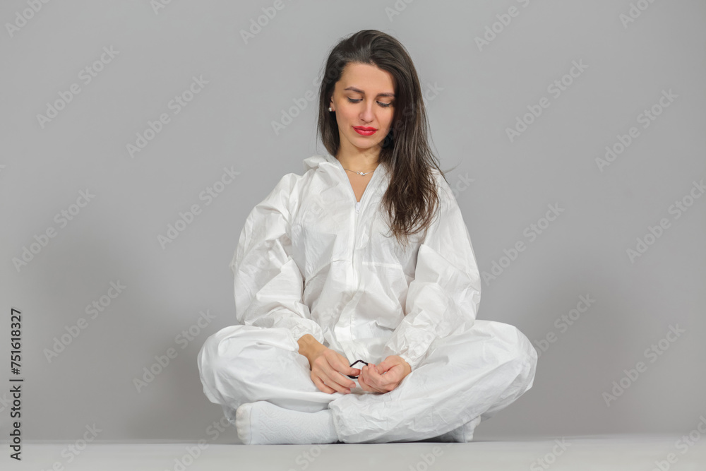 Headshot of woman in white protective suit of synthetic paper, glasses and respiritory half mask, sitting in lotus position, looking down, on gray background