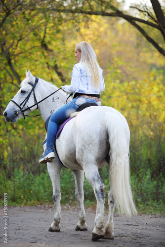 Blonde woman rides on white horse on path in autumn park, back view