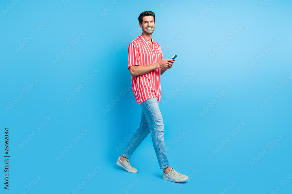Full size photo of busy pleasant man dressed striped shirt denim pants hold smartphone go to empty space isolated on blue color background Full size photo of busy pleasant man dressed striped shirt denim pants hold smartphone go to empty space isolated on blue color background