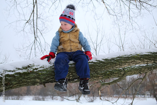 Little happy boy sits on thick branch of tree in winter snowy day