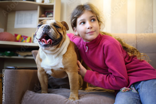 Girl hugging an English bulldog on a couch in the room, focus on the dog photo