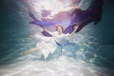 Smiling young woman in white dress poses in swimming pool with dark-blue fabric in hands underwater