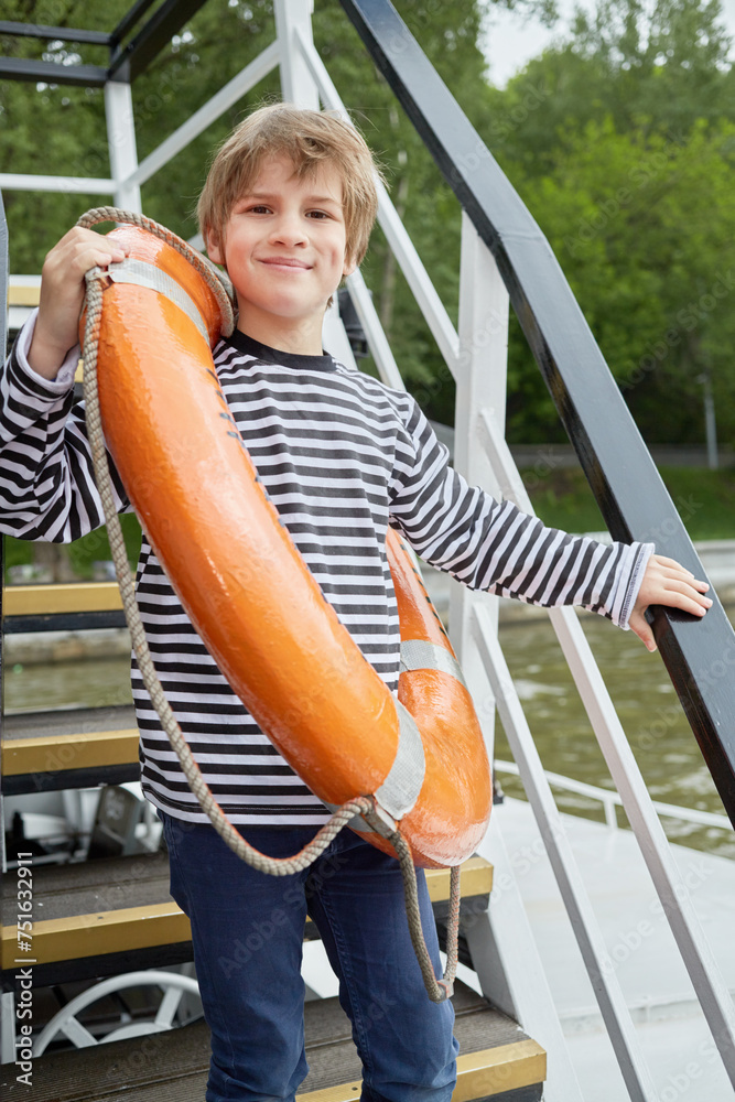 Smiling boy in striped vest with lifebouy ring over shoulder stands on pleasure boat deck Smiling boy in striped vest with lifebouy ring over shoulder stands on pleasure boat deck