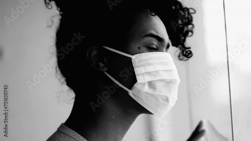 One confined young black woman imprisoned at home during pandemic looking out from apartment window wearing surgical face mask feeling trapped, monochromatic photo
