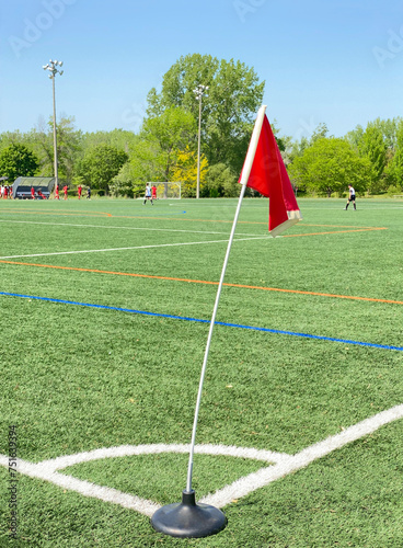 Red flag on the corner of a soccer pitch. Football red flag on the green synthetic grass. Waving flag on the playing field. Players in the background.