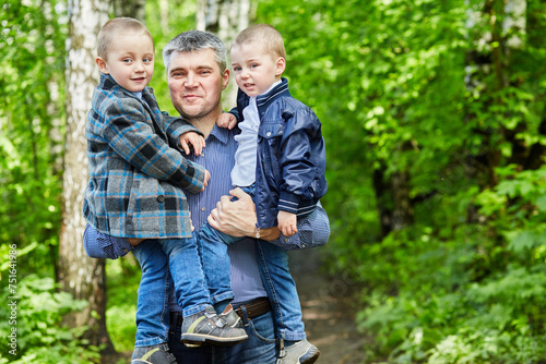 Father holds two sons on his arms standing on pathway in summer park