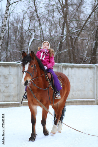 Little girl in winter clothes sitting on horseback at the equestrian site in front of trees
