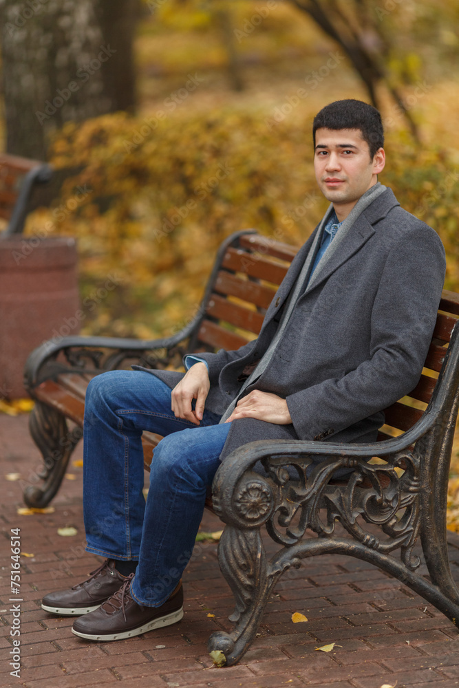 Portrait of young man in gray coat and jeans sitting on bench in alley in park Portrait of young man in gray coat and jeans sitting on bench in alley in park