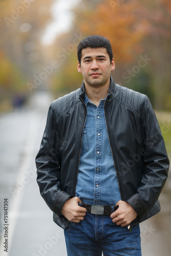 Half length portrait of young dark-haired man in black jacket on avenue in park, hands on his belt