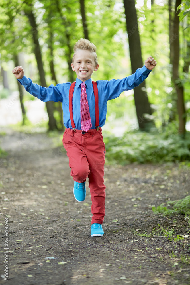 Happy boy in red trousers with braces and blue shirt runs along path in summer park