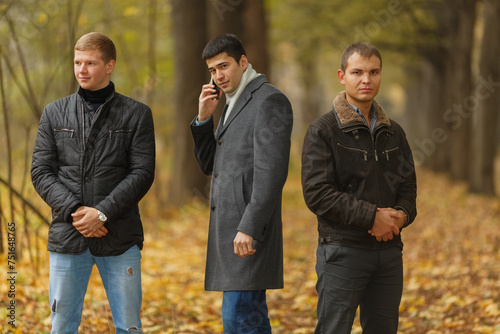 Portrait of three young men in autumn park, one talking on phone, one looking at camera, one looking to side