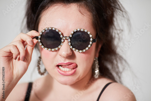 Dark-haired girl model in stylish round dark glasses shows tongue posing in studio