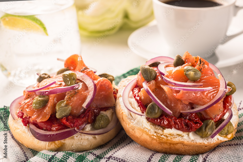 Two sandwiches with cheese cream, red fish, onions and capers on a napkin against the background of a cup of black coffee close-up. Two sandwiches with cheese cream, red fish, onions and capers on a napkin against the background of a cup of black coffee close-up.