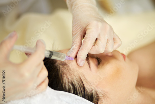 Beautician hands in rubber gloves makes injection into above eyebrow area to woman lying on couch in beauty salon.