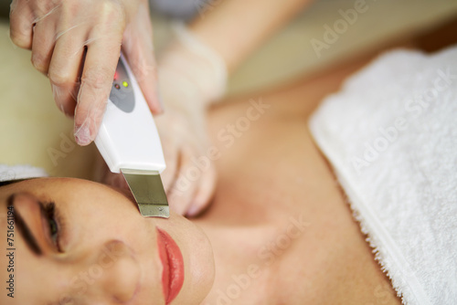 Beautician hands in rubber gloves do ultrasonic skin cleaning on face of woman lying on couch in beauty salon.
