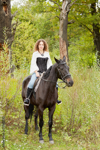 Smiling woman with curly hair sits on bay horse in the park.