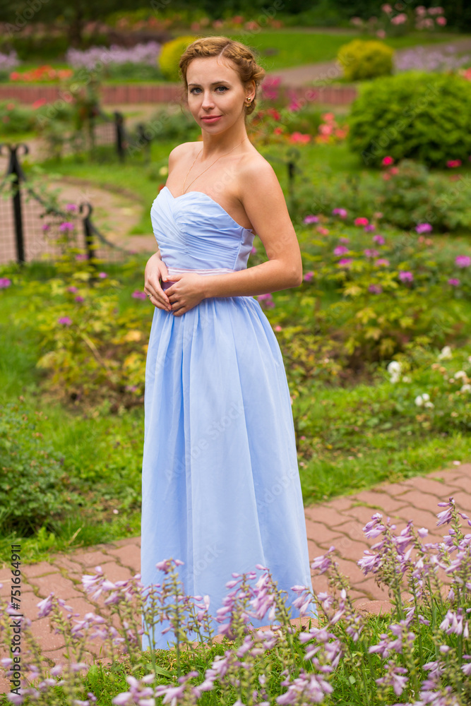 Pretty woman in long blue dress stands near flowers in summer garden Pretty woman in long blue dress stands near flowers in summer garden