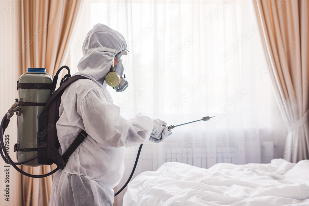 Faceless pest control worker in a protective suit sprays insect poison in bedroom. White room.