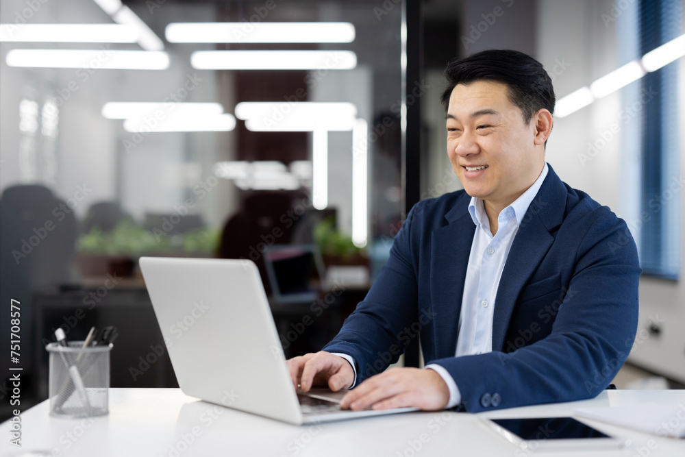 Successful and smiling young Indian man sitting in a modern office at a desk in a suit and working on a laptop, texting and typing on a keyboard. Successful and smiling young Indian man sitting in a modern office at a desk in a suit and working on a laptop, texting and typing on a keyboard.