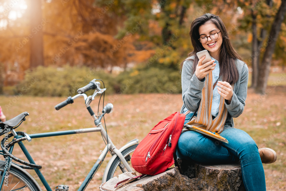 Obraz premium Young female student using mobile phone while studying for exam in public park during the sunny Autumn day.