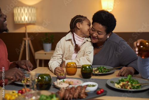 Portrait of African American little girl kissing grandma on cheek at dinner table with family copy space 