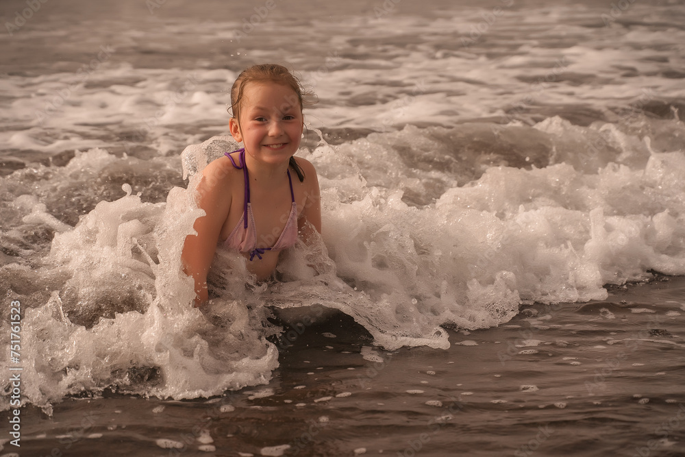 A happy young girl in a purple swimsuit enjoys the foamy sea waves on a sunny day. Joyful Child Playing in Sea Waves