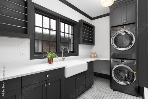 A laundry room with dark grey cabinets, grey over under washer and dryer, farmhouse sink, pattern tile floor, and white marble countertops. No brands or labels.