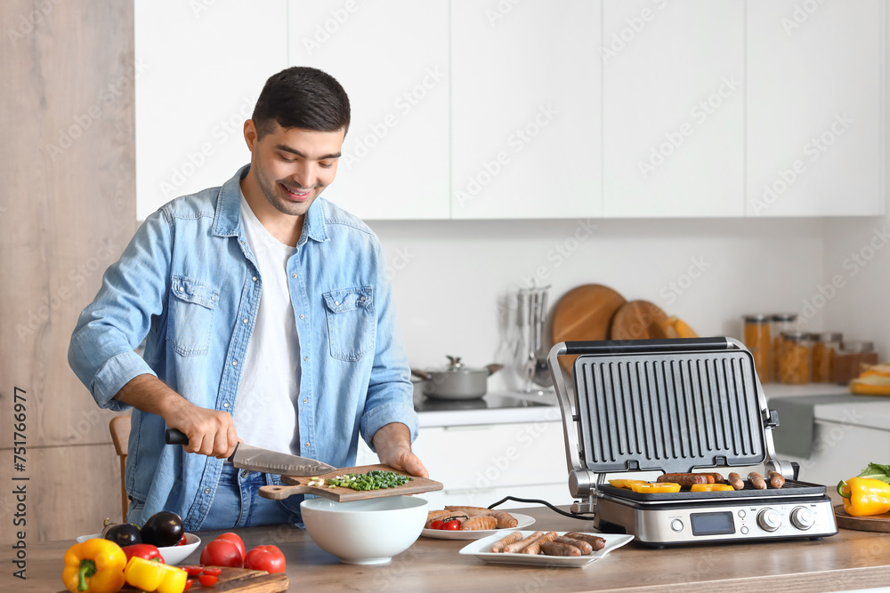Young man cooking tasty sausages on modern electric grill and making salad of fresh vegetables in kitchen Young man cooking tasty sausages on modern electric grill and making salad of fresh vegetables in kitchen