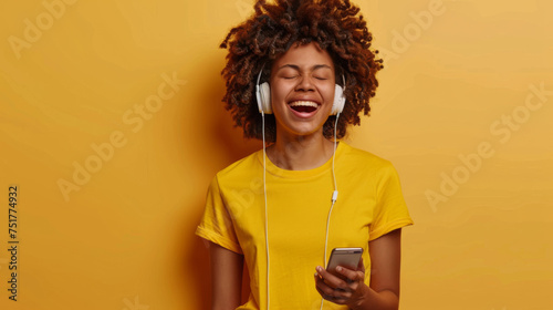 A joyful young woman with closed eyes sings while listening to music on her smartphone with headphones, against a vibrant yellow background.