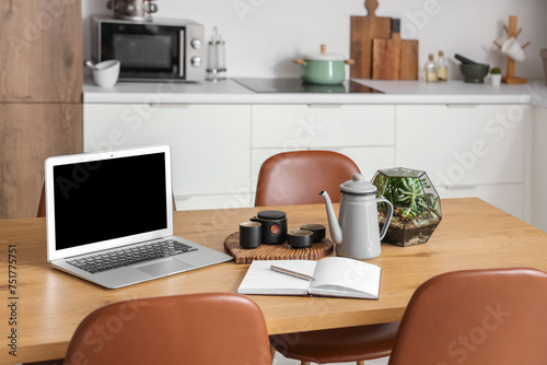Laptop with blank screen, tea set and notebook on table in modern kitchen © Pixel-Shot