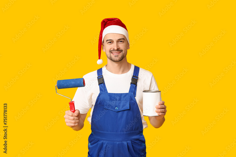 Portrait of male painter in Santa hat with roller and can of paint on yellow background Portrait of male painter in Santa hat with roller and can of paint on yellow background