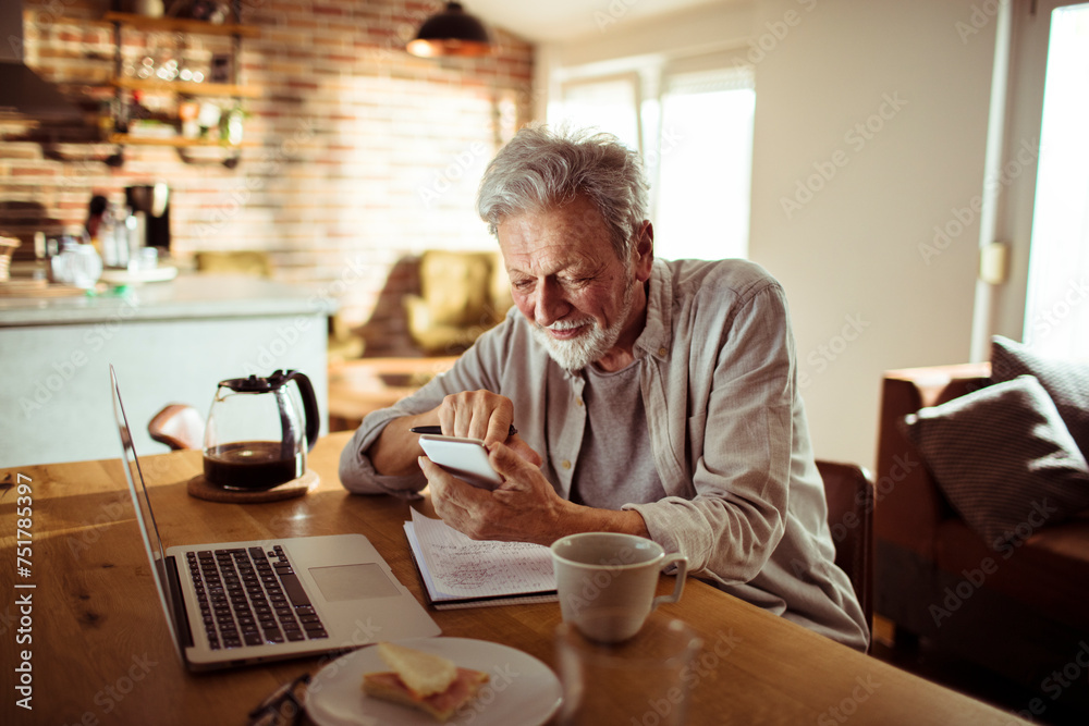 Smiling senior man using smartphone with laptop on desk at home Smiling senior man using smartphone with laptop on desk at home