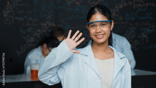 Cute girl waving hands to camera while people doing experiment at laboratory. Young cute student standing blackboard with chemical theory with blurring background at STEM science class. Edification.