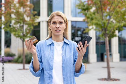 Blue eyed woman standing next to shopping mall with debit card and smartphone in raised hands. Stressed female feeling frustrated because of uncharged device and inability checking bank balance. photo