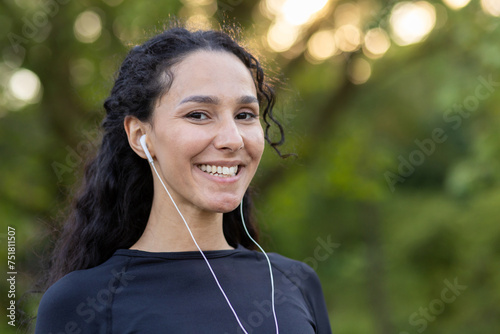 Optimistic diverse sportswoman in black top smiling at camera while listening to motivating playlist in wired earphones. Attractive female in black top finishing training in green public area. © Liubomir