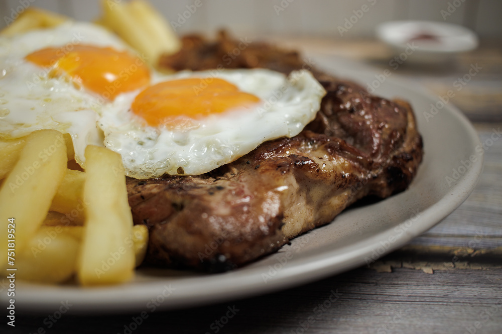 Bistec a lo pobre. Beef, eggs and fried potatoes on a plate on a wooden table with cutlery. Close-up. Typical Chilean food
