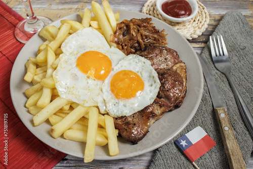 Bistec a lo pobre, beef, eggs, and French fries on a plate on a wooden table with cutlery. Typical Chilean food. Aerial view with a glass and Chilean flag