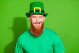 Photo of flirty cool man with ginger beard dressed green t-shirt headwear blinking eye on partric day isolated on green color background