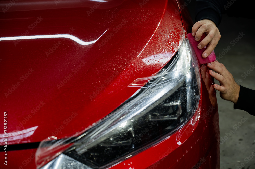 The master applies vinyl film to the headlight of a red car. Closeup view on worker detailer hand smoothing with a scraper protective film. 