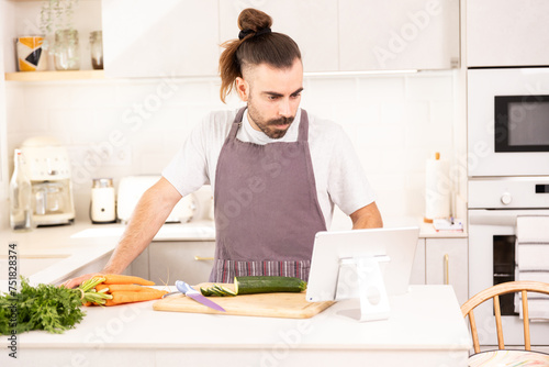 long-haired man cooking at home looking at tablet, chopping vegetables