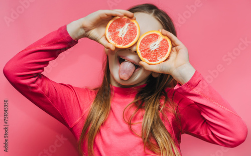 Little cute funny girl using grapefruit instead of eyes, having fun and fooling around in the studio on a pink background and showing her tongue. Concept of citrus diet, fruits and vitamins.