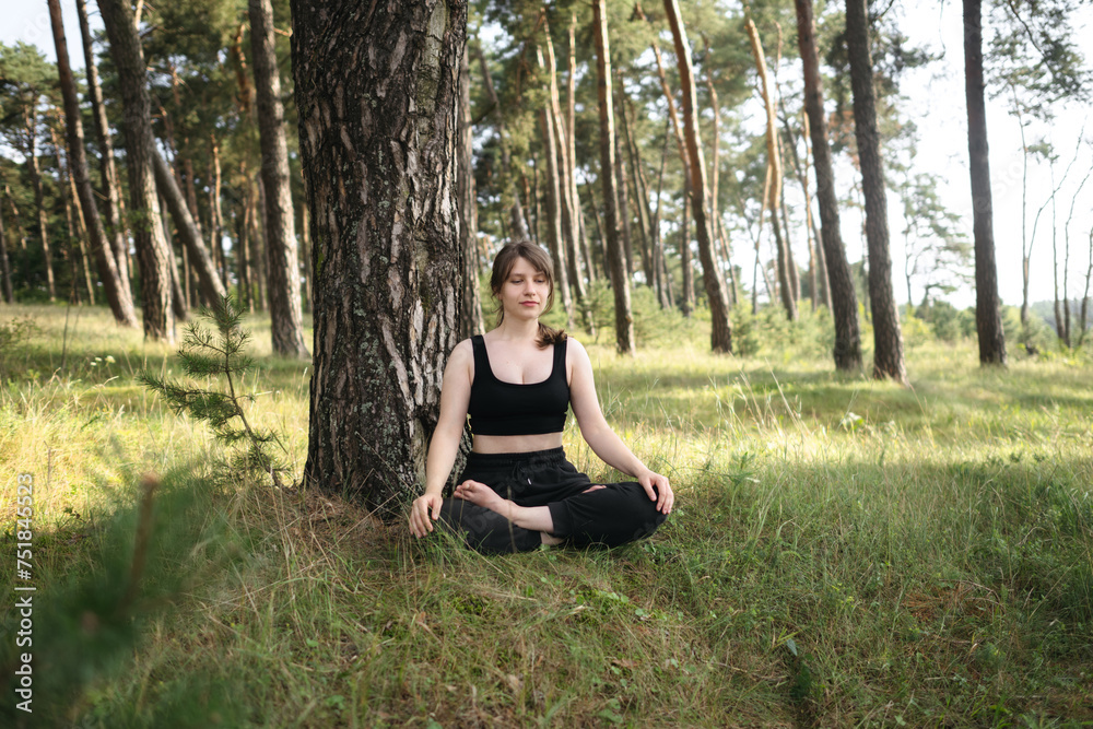 Young woman practices yoga and meditates in lotus position in the pine forest. Young attractive girl relaxing and doing exercises in nature. Girl resting after training. Exercise, woods, sun, summer. Young woman practices yoga and meditates in lotus position in the pine forest. Young attractive girl relaxing and doing exercises in nature. Girl resting after training. Exercise, woods, sun, summer.