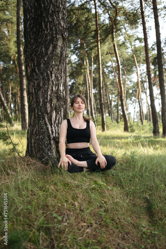 Young woman practices yoga and meditates in lotus position in the pine forest. Young attractive girl relaxing and doing exercises in nature. Girl resting after training. Exercise, woods, sun, summer.