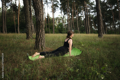 Yoga cobra pose by woman in black suit on green grass in the park around pine trees. Alone yoga in the garden in the morning, stretch back. Healthy lifestyle. 