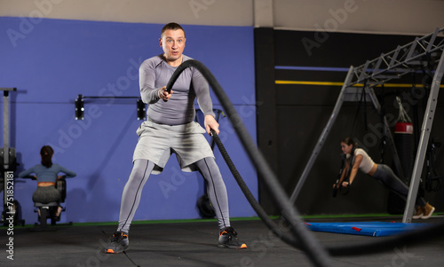 Adult athletic man in sportswear training with rope in gym