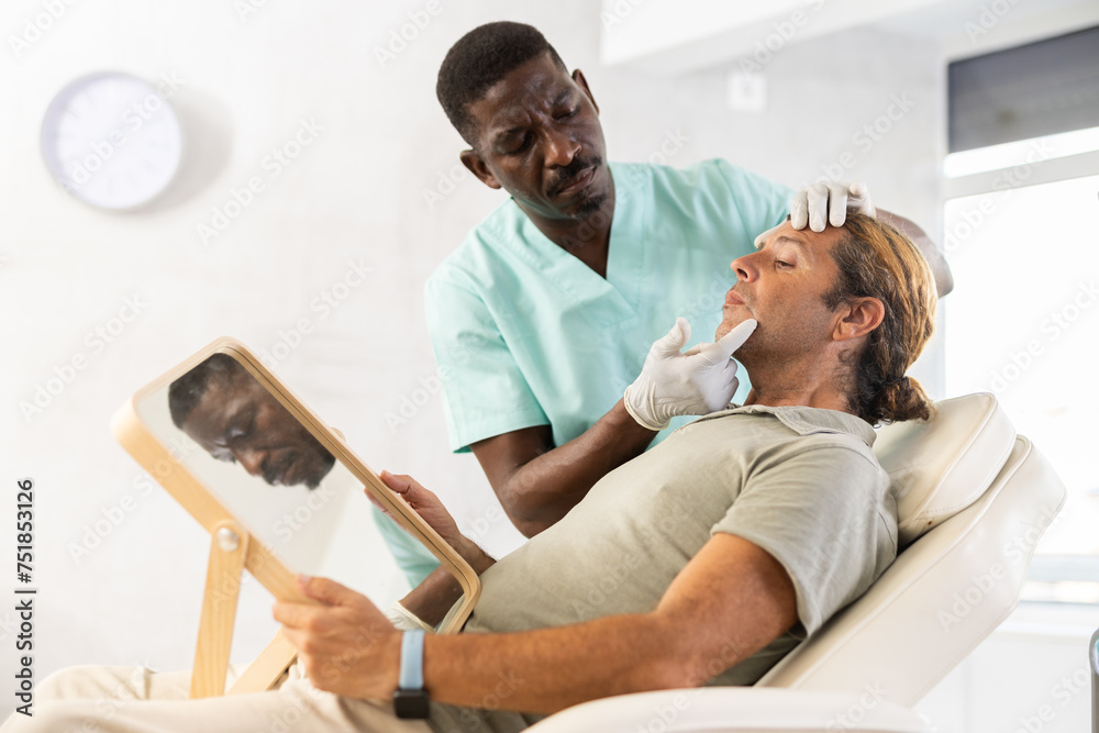 Experienced African male cosmetologist examines facial skin of man patient and advises on possibility of rejuvenation procedure.