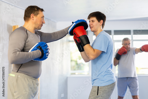 Concentrated sporty young guy in boxing gloves practicing self-defense techniques in gym, throwing punches on focus mitts in hands of instructor.. © JackF