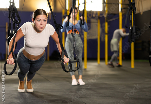 Slender athletic girl work out muscles of arms and shoulders on TRX rope loop simulator in gym of fitness club. Gym visitor trains triceps during classes at multi-station in wellness center.