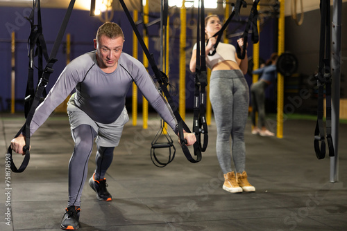 Adult male athlete with group working out on gymnastics rings at modern crossfit gym